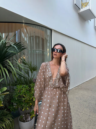 Woman in a brown dress and cap standing on a sunlit outdoor patio with palm trees and buildings in the background.